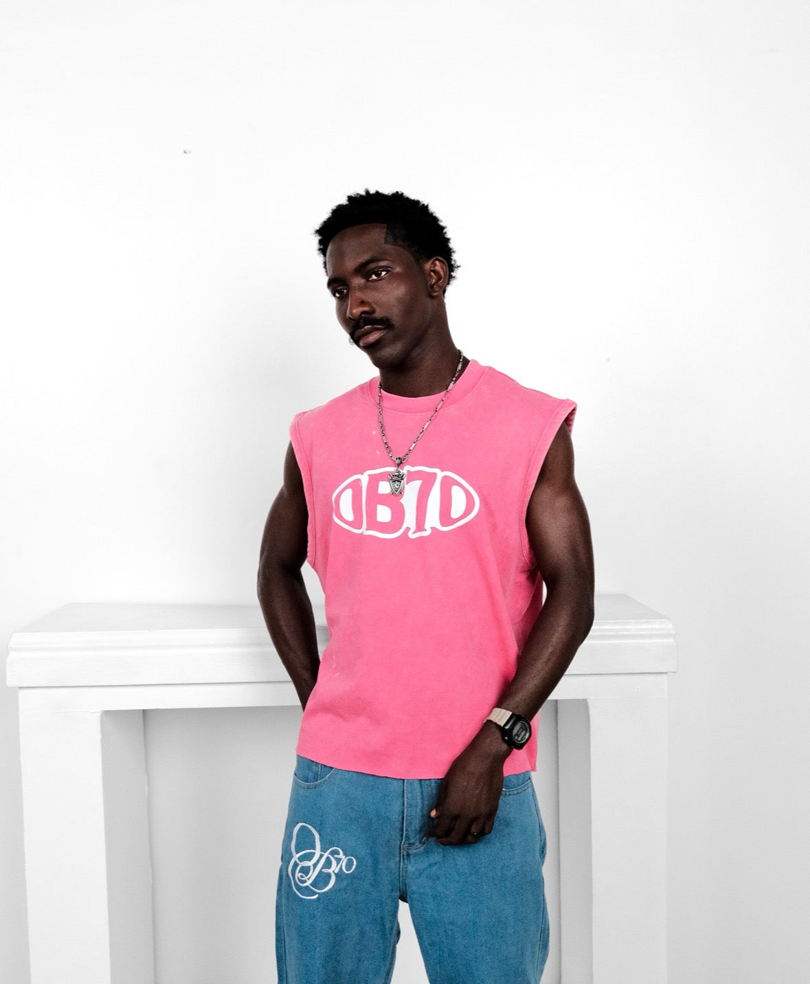 Man wearing a pink sleeveless shirt with an OB70 logo and blue jeans against a white background