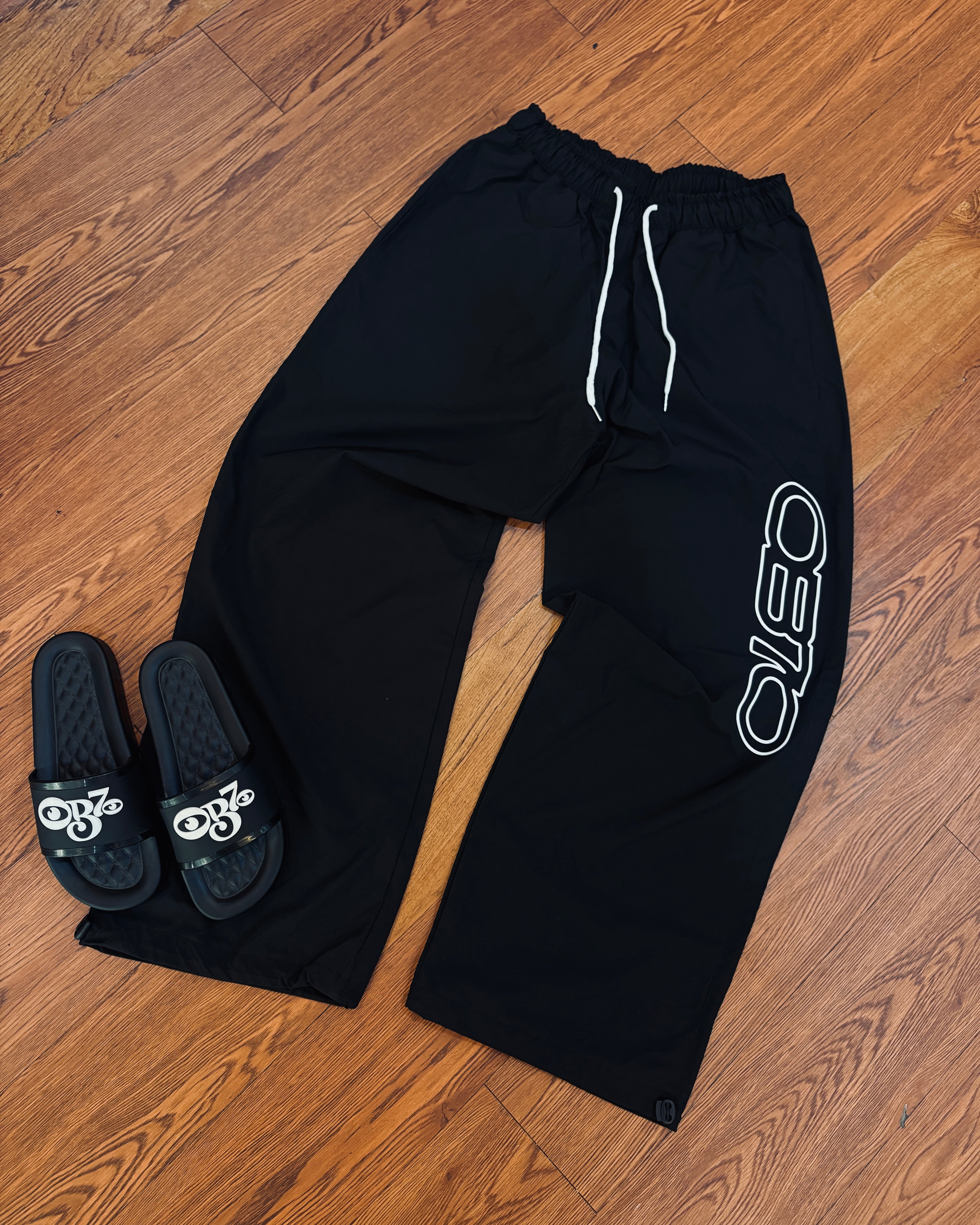 Black sweatpants with a logo and black slides with a logo on a wooden floor.