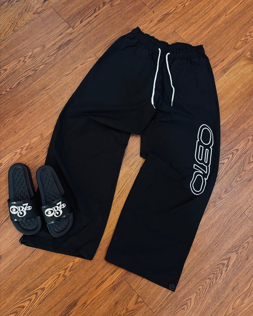 Black sweatpants with a logo and black slides with a logo on a wooden floor.