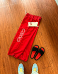 Red pants with a brand logo on a wooden floor, accompanied by black sandals and blue shoes.
