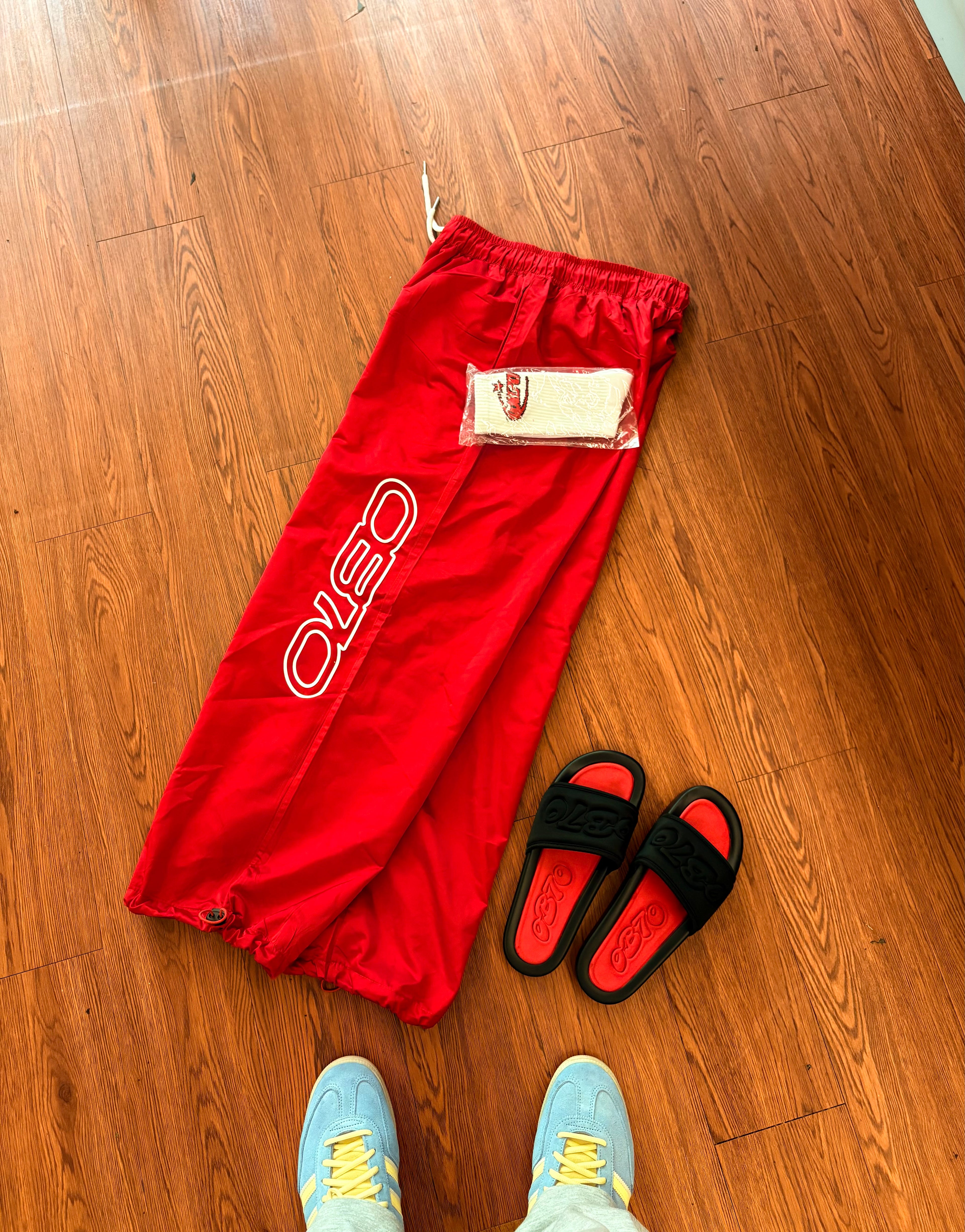 Red pants with a brand logo on a wooden floor, accompanied by black sandals and blue shoes.