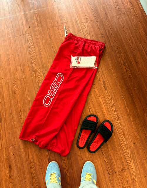 Red pants with a brand logo on a wooden floor, accompanied by black sandals and blue shoes.