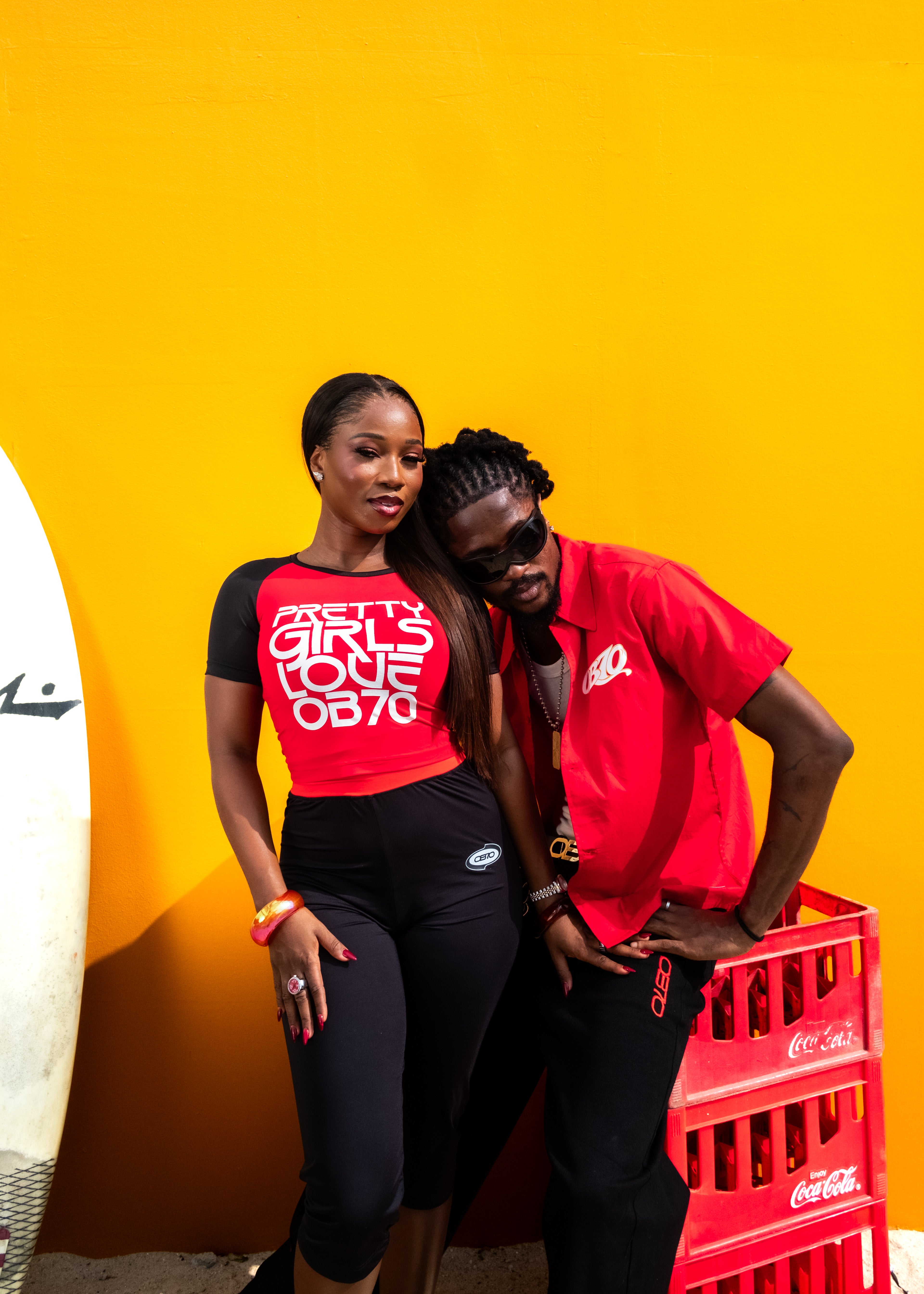 A man and a woman posing together in front of a bright yellow wall. The woman wears a red and black graphic t-shirt with the text 'GIRL'S LOVE OB70’ and black leggings, while the man wears OB70 red short-sleeve button-up shirt and black pants.