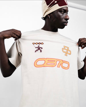 Person wearing a beige OB70 '25 Championship T-shirt, holding it in front of a white background