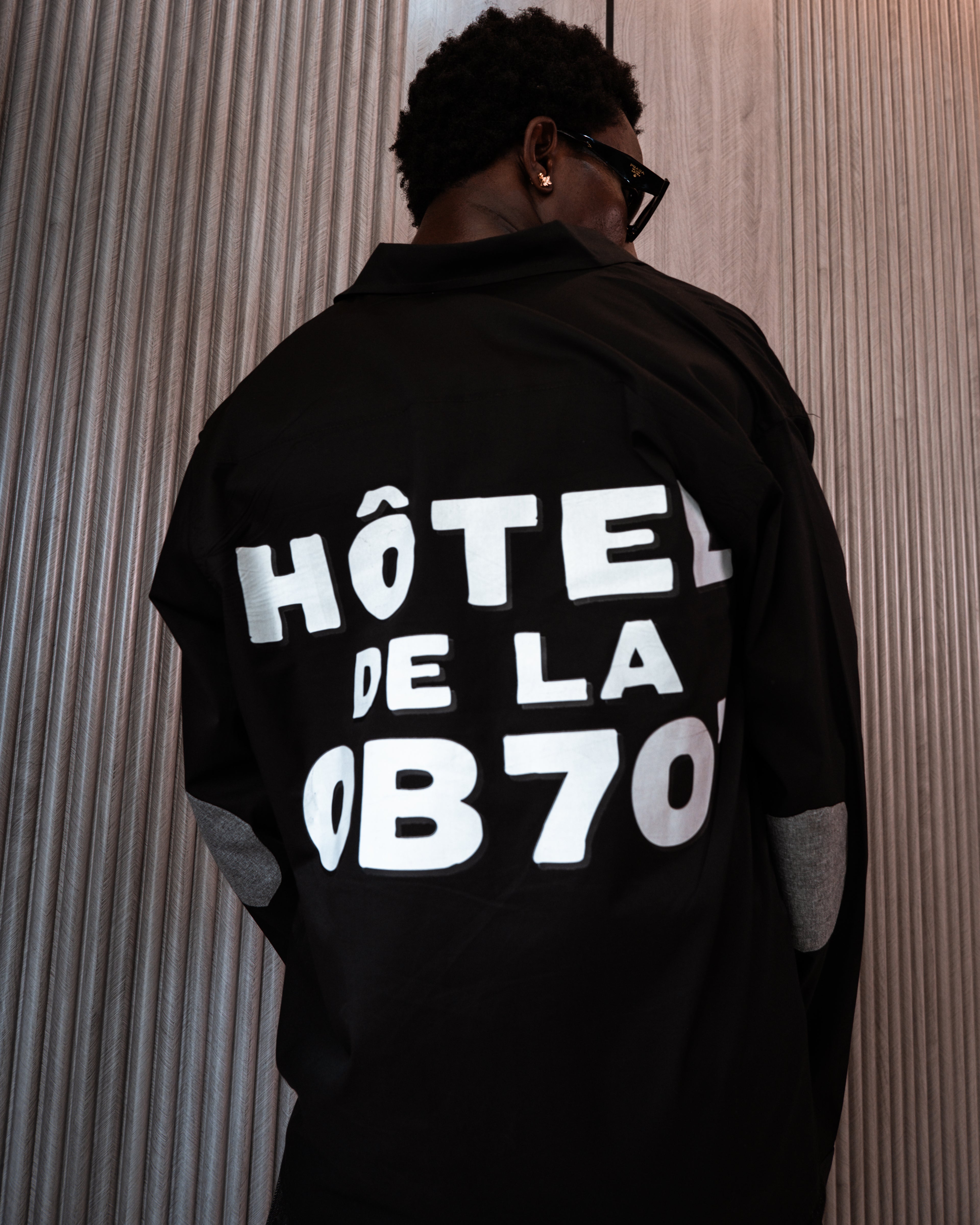 Person wearing a black jacket with 'HôTEL DE LA OB70' text on the back against a striped wall.