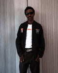 Man wearing a black jacket with logos, white shirt, and dark pants against a textured wall.