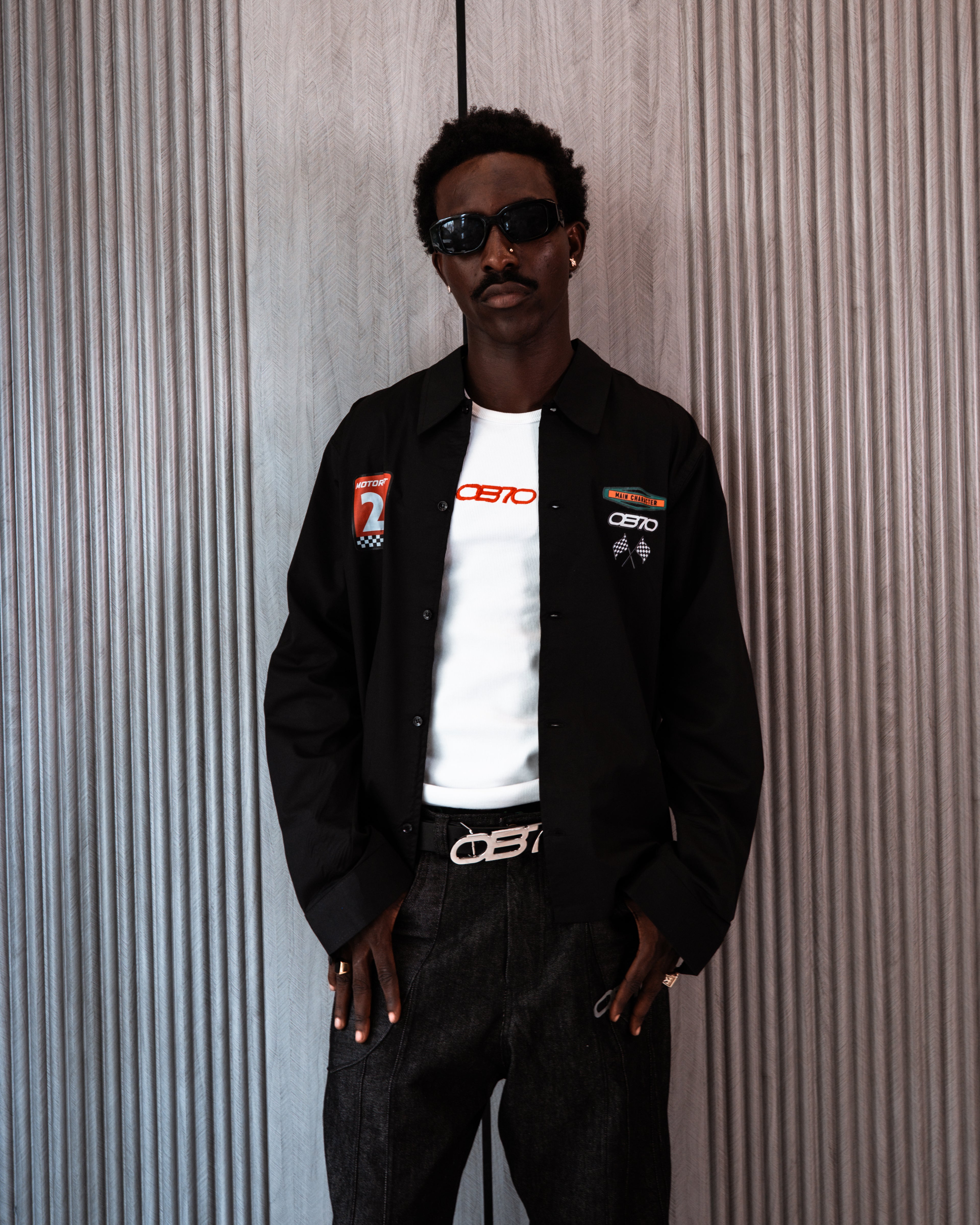 Man wearing a black jacket with logos, white shirt, and dark pants against a textured wall.