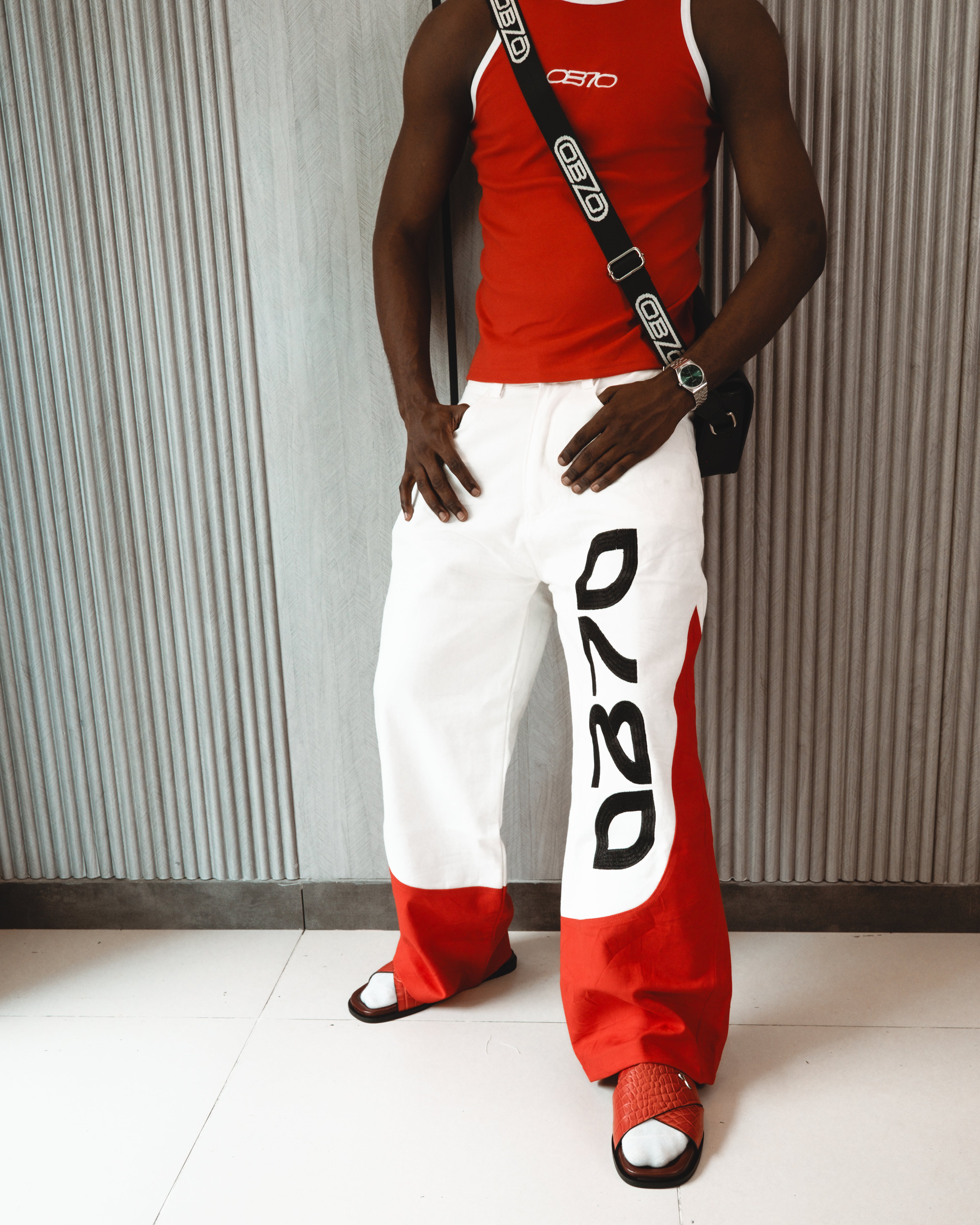 Person wearing a red tank top and white pants with black text, standing against a striped wall.
