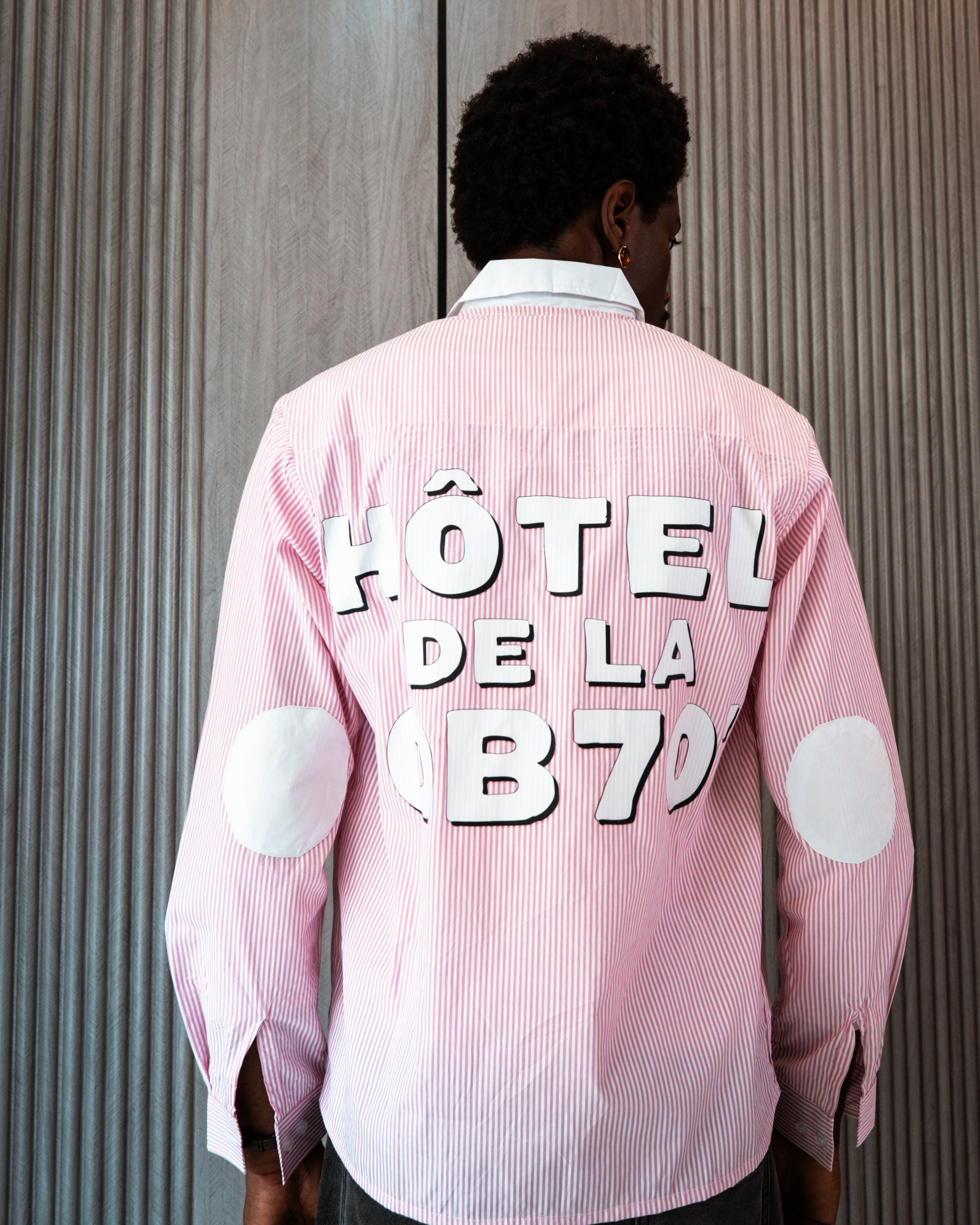 Person wearing a pink shirt with 'HôTEL DE LA OB70' text on the back against a striped wall.