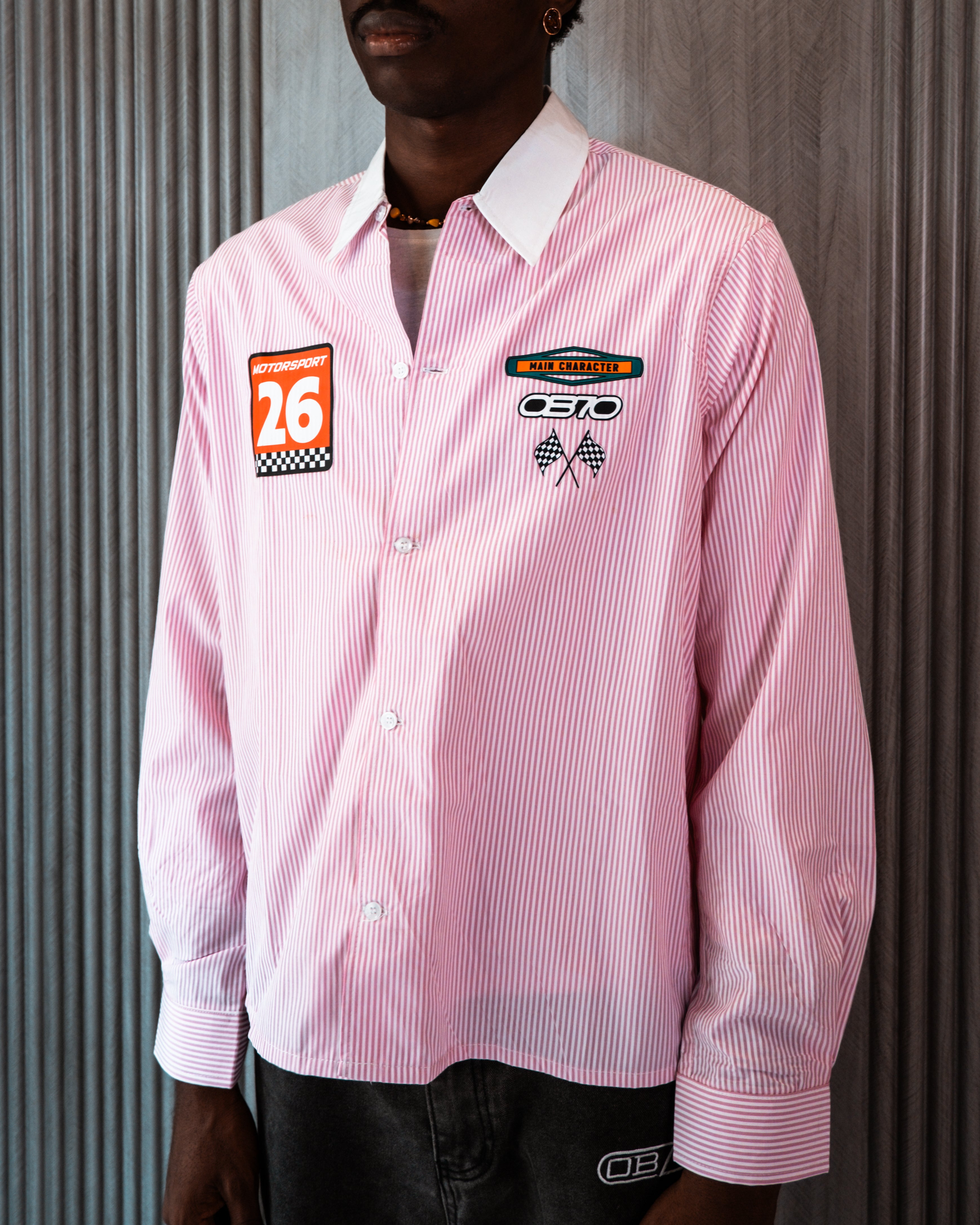 Person wearing a pink striped shirt with racing-themed patches against a corrugated metal background